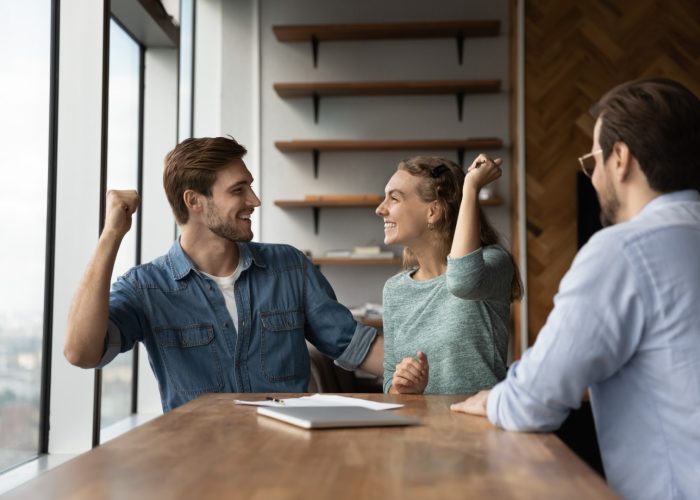 Happy excited couple feeling joy in realtor office, celebrating getting mortgage, renting or buying house for living together, making winner gesture, laughing. Real estate, property purchase concept